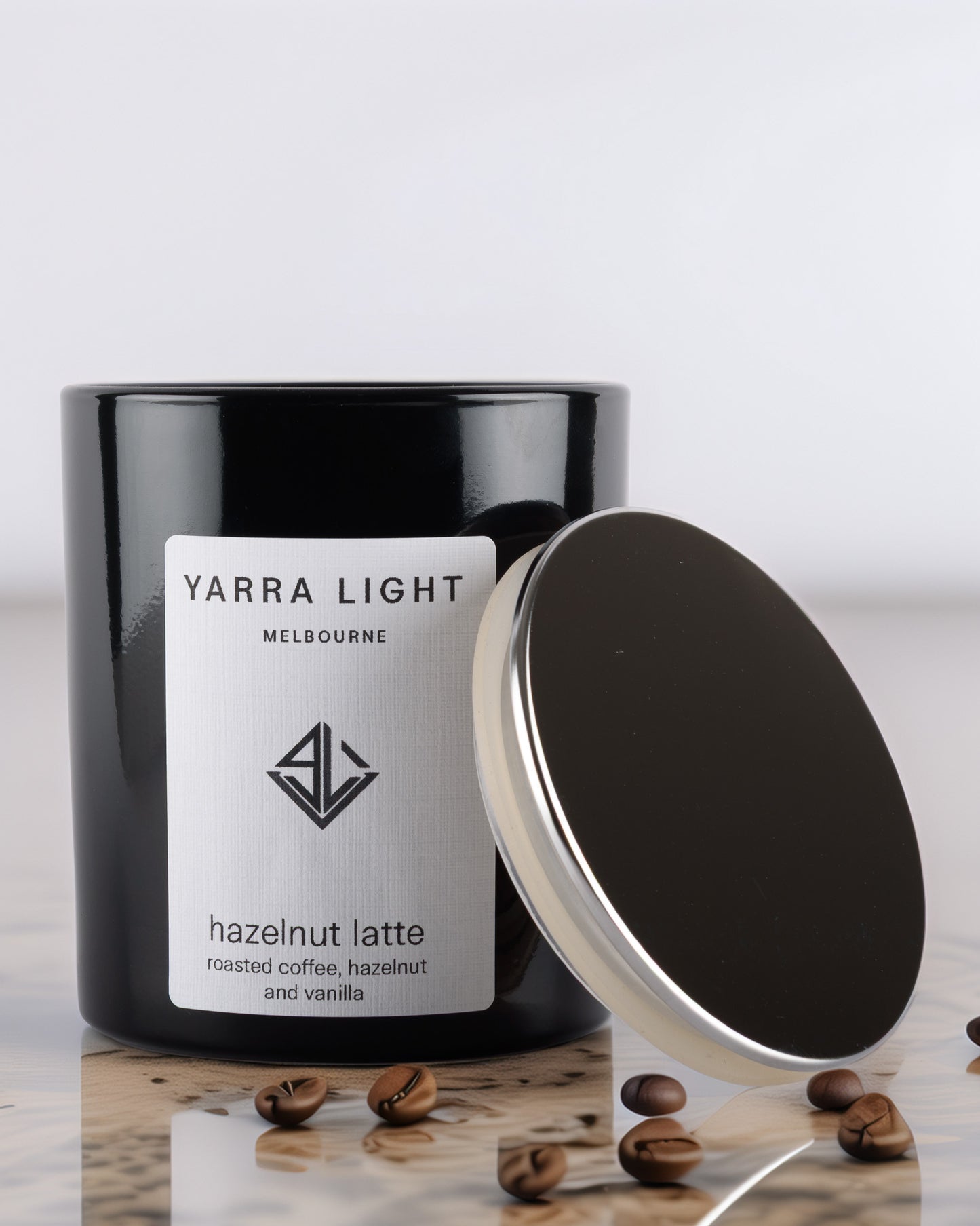 Black glossy soy wax candle labeled 'Yarra Light' with hazelnut latte scent on a reflective surface .
