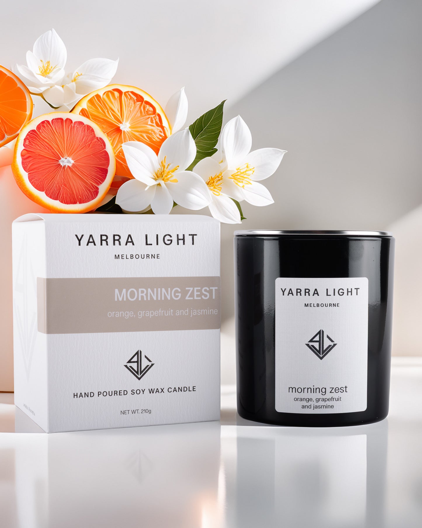Yarra Light Morning Zest candle and packaging on a reflective surface with flowers and citrus fruits in the background.