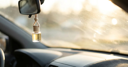 A small glass car diffuser bottle with a timber cap hanging from a rear-view mirror in warm afternoon sun