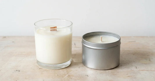 Two soy candles side by side on a bench, one in clear glass and one in a metal tin, unlit