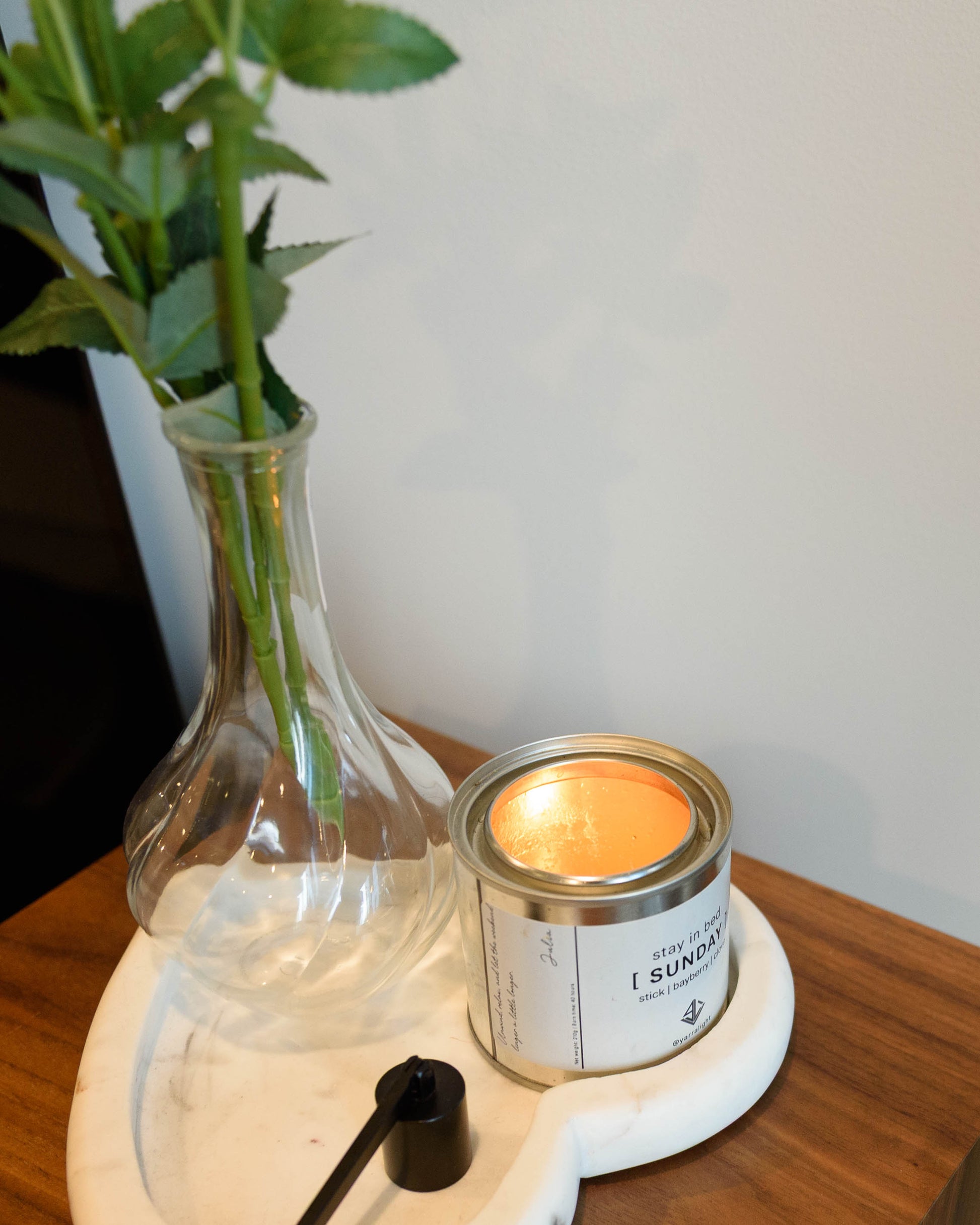 Candle in a can with a label next to a clear vase with green stems on a wooden surface.