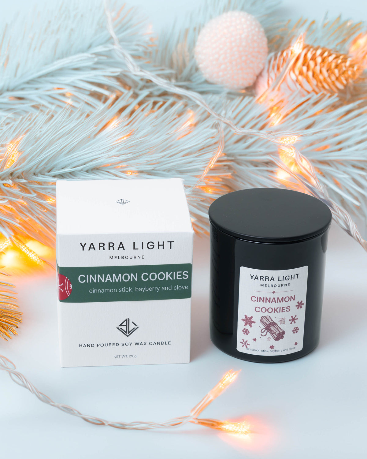 Cinnamon Cookies Christmas soy candle in a black glass jar beside its gift box, with frosted pine branches and fairy lights