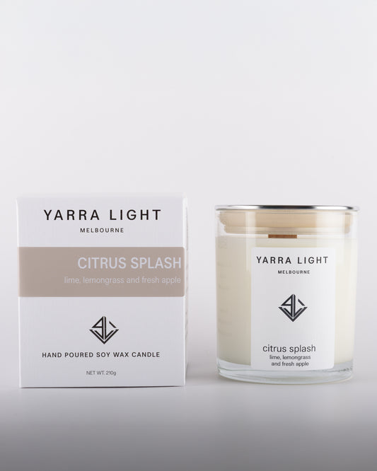 Citrus Splash soy wax scented candle with wood wick in a clear glass jar with minimalist label, placed beside its eco-friendly packaging box in warm neutral tones, inspired by citrusy notes of lime and lemongrass.