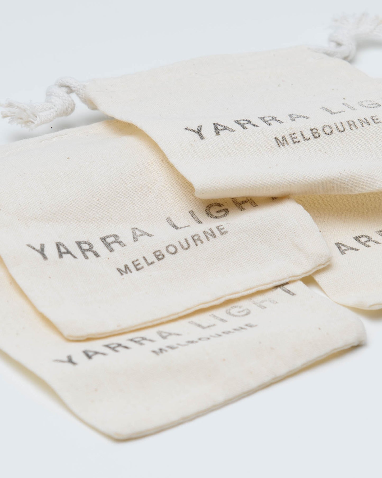 Four small cream-colored fabric drawstring pouches stacked on a white background, each printed with the text 'YARRA LIGHT MELBOURNE' in a dark gray serif font.