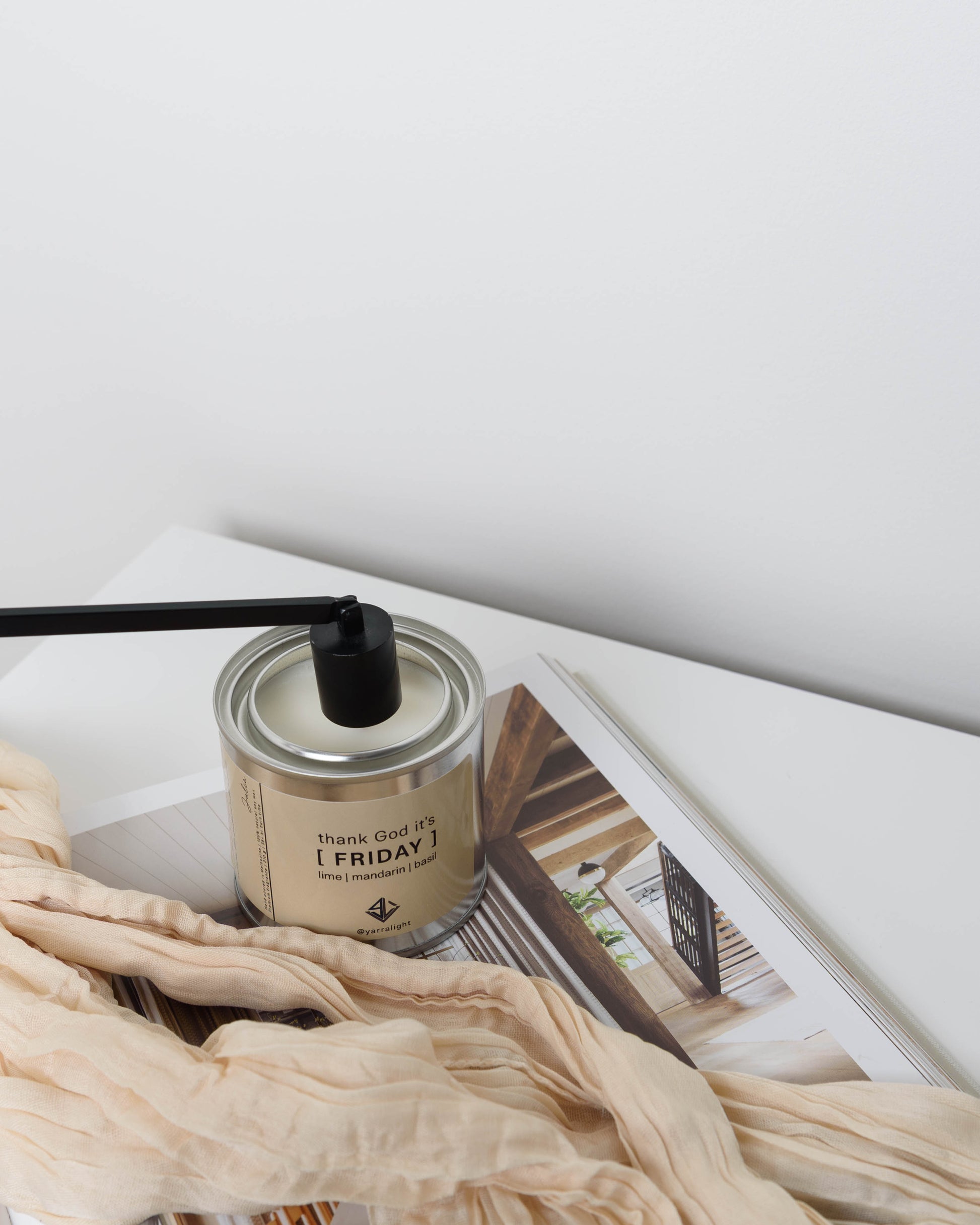 Yarra Light scented candle with a black snuffer and a cream-colored label reading 'THANK GOD IT'S [FRIDAY] is styled on top of an open magazine with a beige silk scarf draped across the foreground against a clean white background.