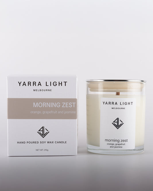 Morning Zest soy wax scented candle with wood wick in a clear glass jar with minimalist label, placed beside its eco-friendly packaging box in warm neutral tones, inspired by citrusy notes of orange and grapefruit.