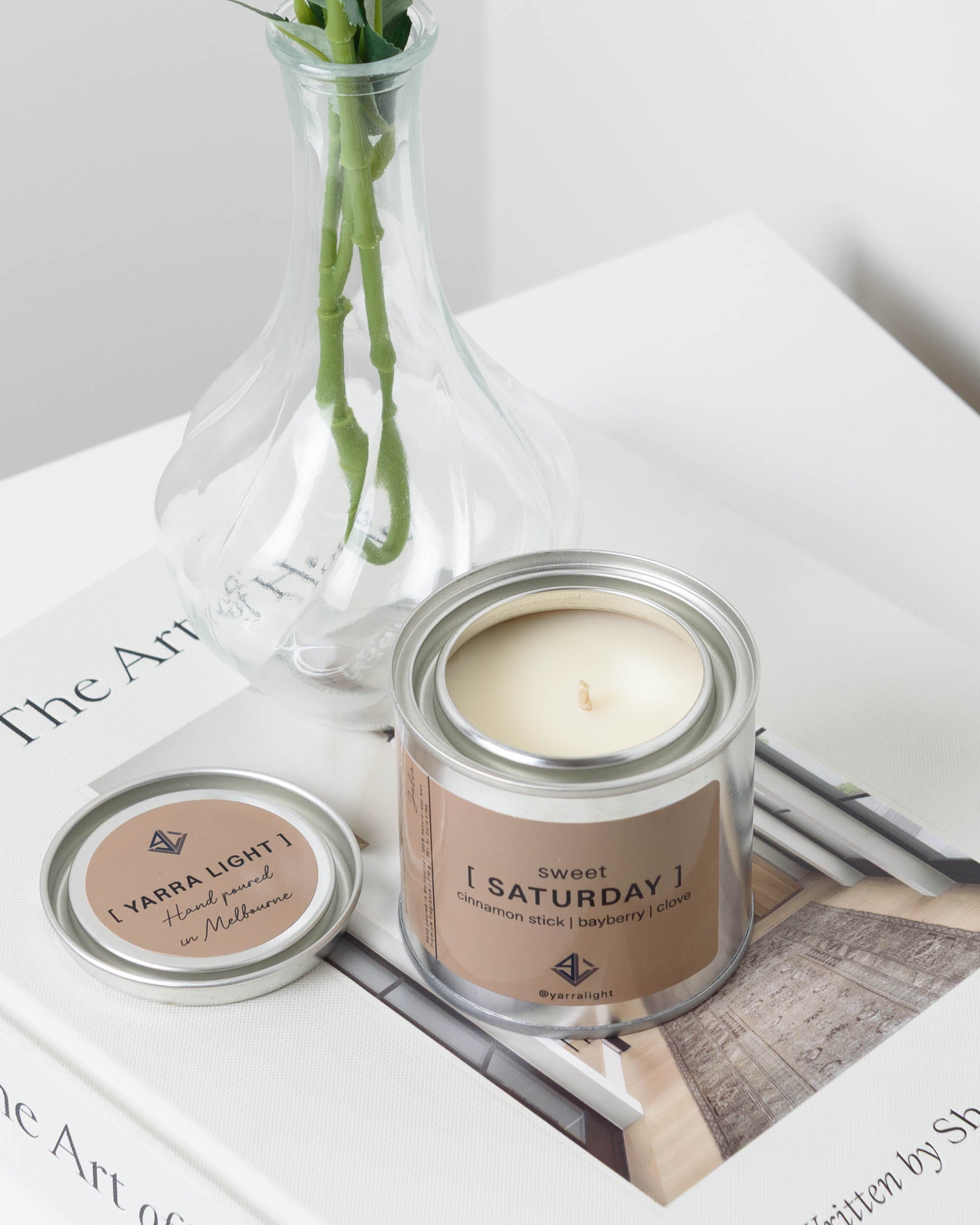 Candle in a metal container labeled 'Sweet Saturday' on a magazine in the background.