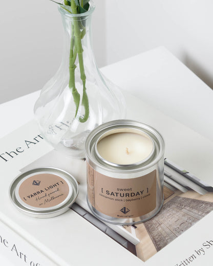 Candle in a metal container labeled 'Sweet Saturday' on a magazine in the background.