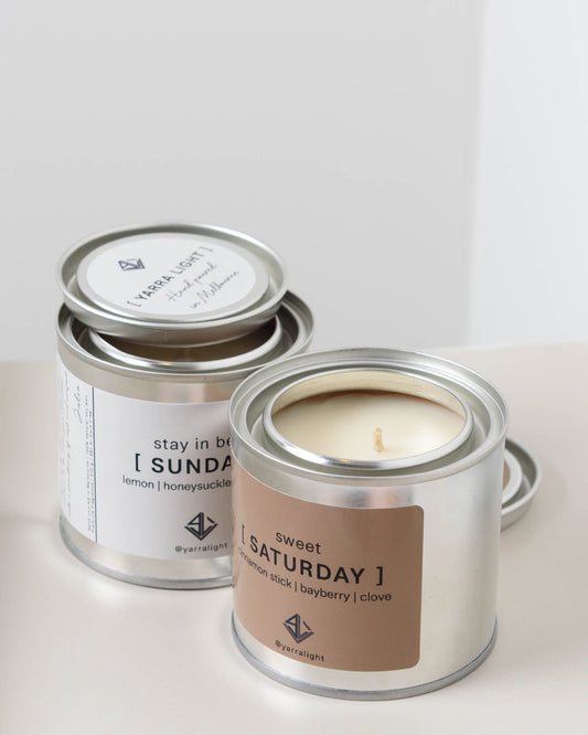 Two candles in metal containers labeled 'Sunday' and 'Saturday' on a light background.