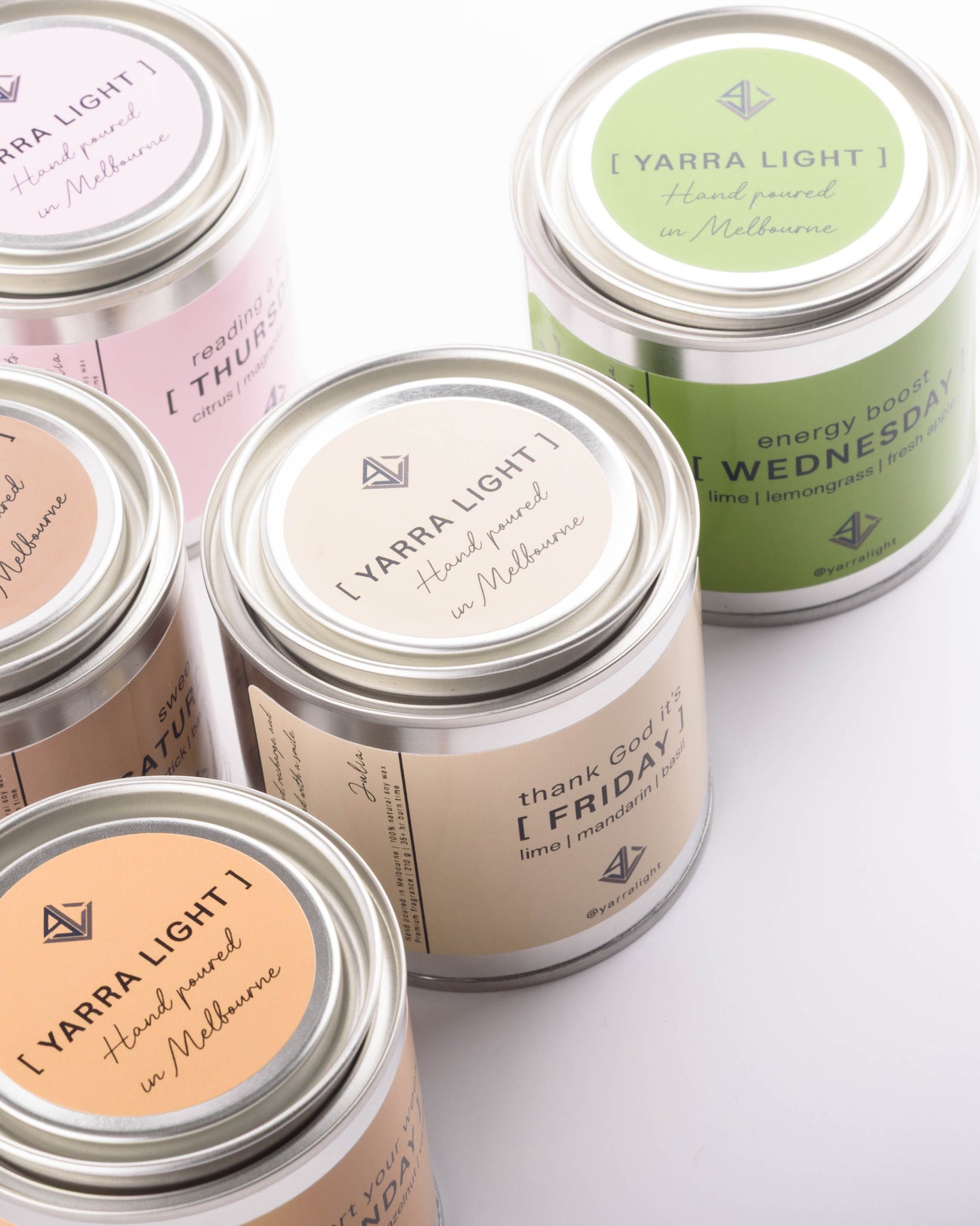 Several metal tin candles with silver lids are arranged on a white background. The tins have pastel labels in pink, beige, peach, and green, each printed with the text '[ YARRA LIGHT ]' and handwritten-style day names such as Thursday, Wednesday, Friday, and Sunday.