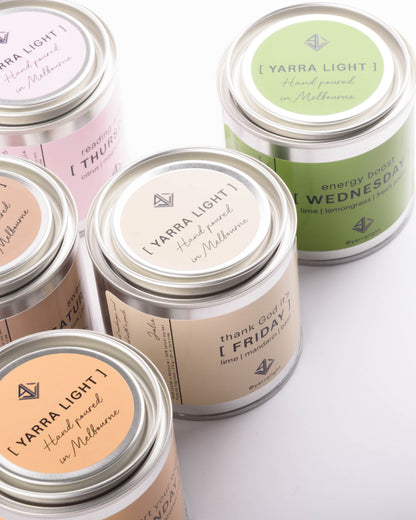 Several metal tin candles with silver lids are arranged on a white background. The tins have pastel labels in pink, beige, peach, and green, each printed with the text '[ YARRA LIGHT ]' and handwritten-style day names such as Thursday, Wednesday, Friday, and Sunday.