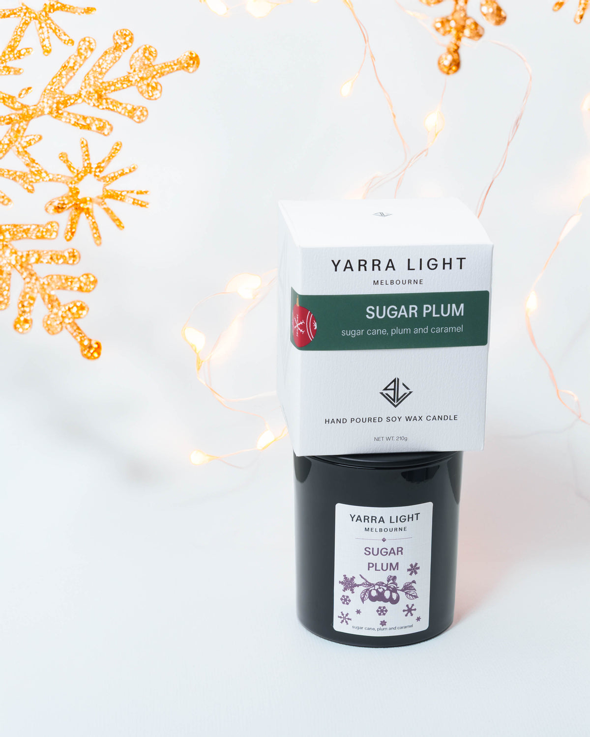 Sugar Plum Christmas soy candle in black glass with its gift box stacked on top, gold snowflakes and fairy lights