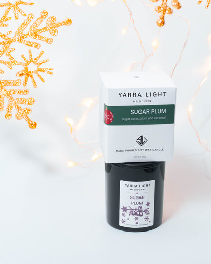 Sugar Plum Christmas soy candle in black glass with its gift box stacked on top, gold snowflakes and fairy lights