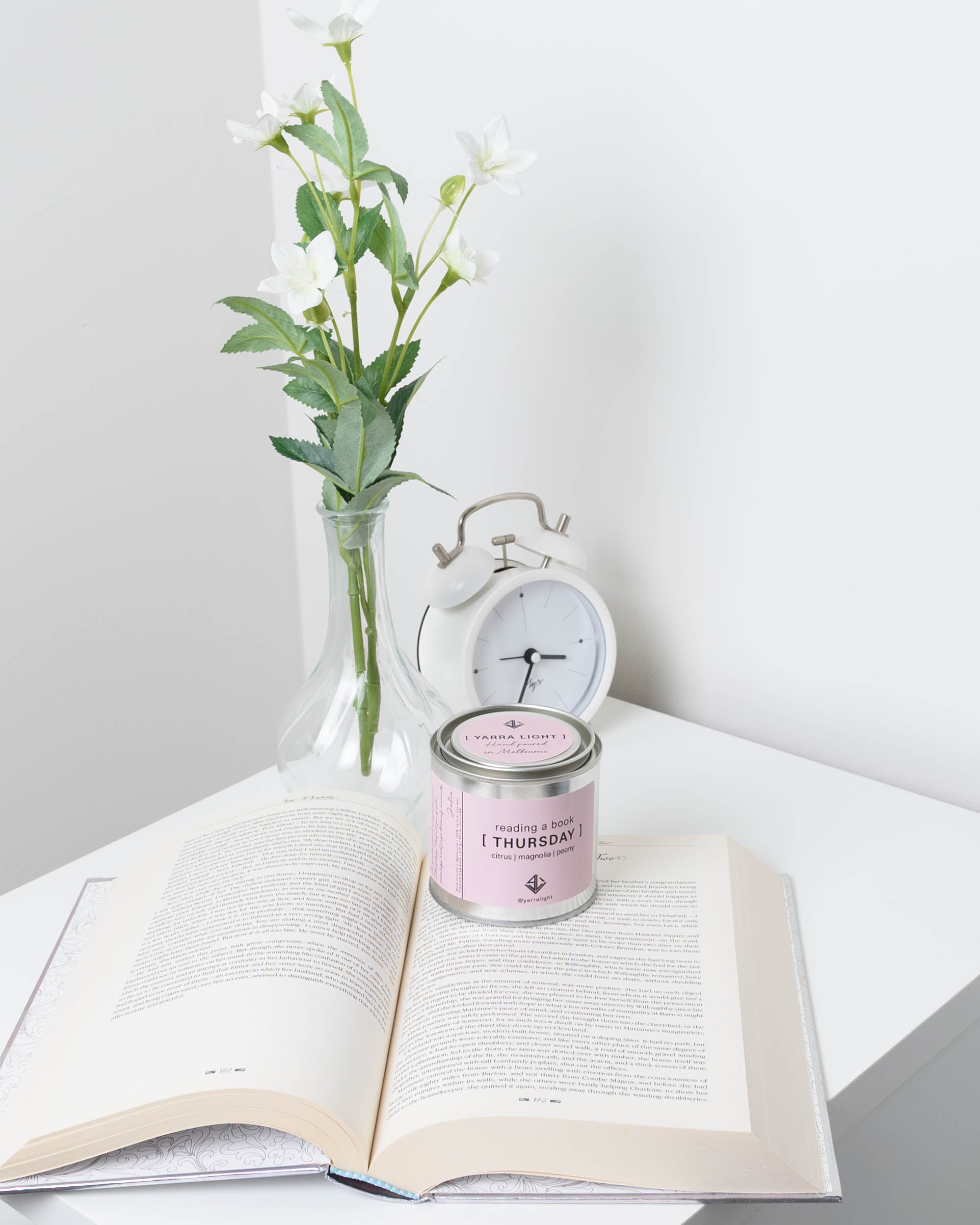 Candle labeled 'Thursday' on an open book with a vase of flowers and alarm clock in the background.
