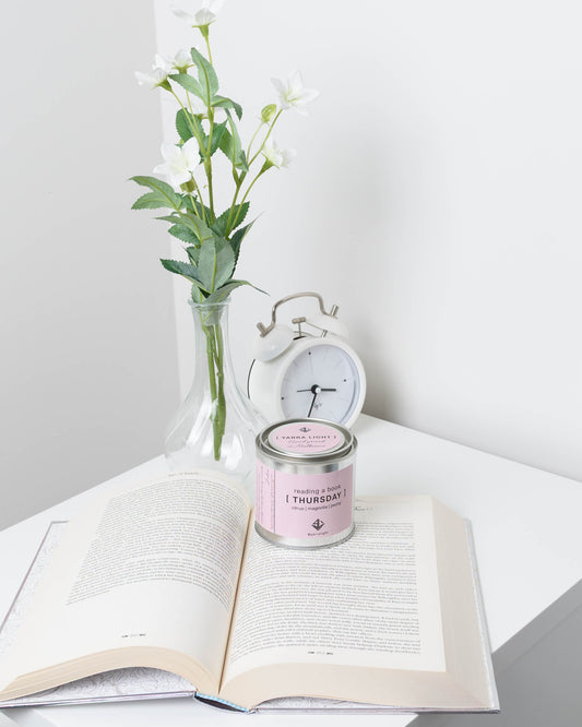 Candle labeled 'Thursday' on an open book with a vase of flowers and alarm clock in the background.