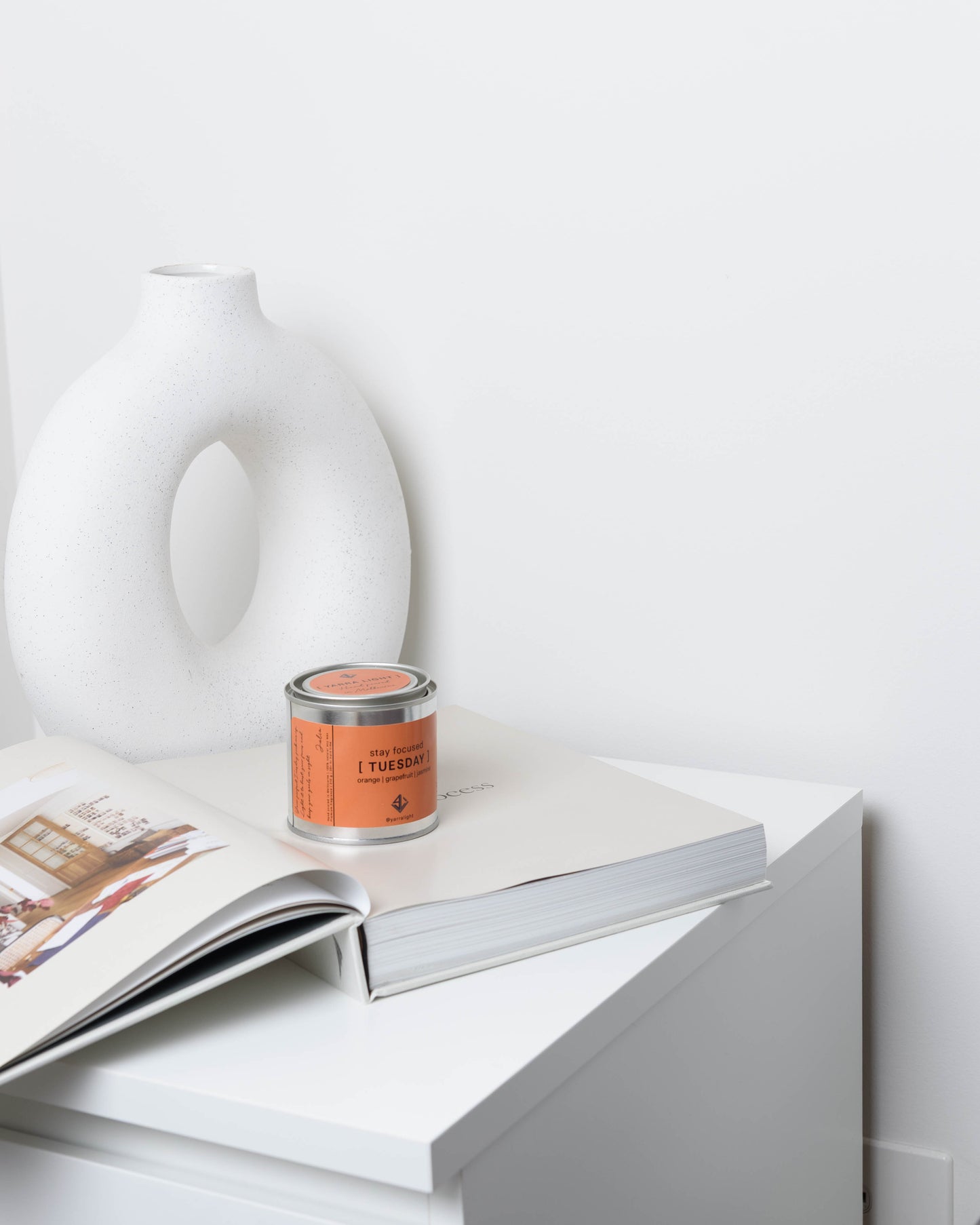 Candle with orange label on a book next to a white vase against a white background